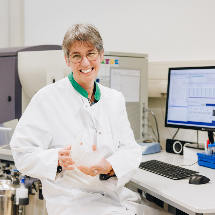 Researcher sits next to a piece of lab equipment and laughingly shows a well plate.