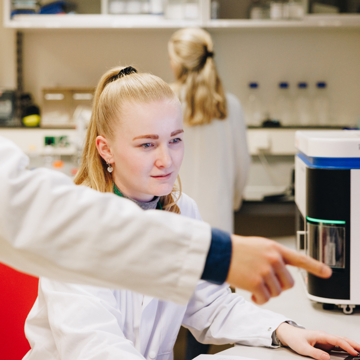 Researcher pointing to something to the right of the image, with other researcher looking. In the background, another researcher is working in a lab